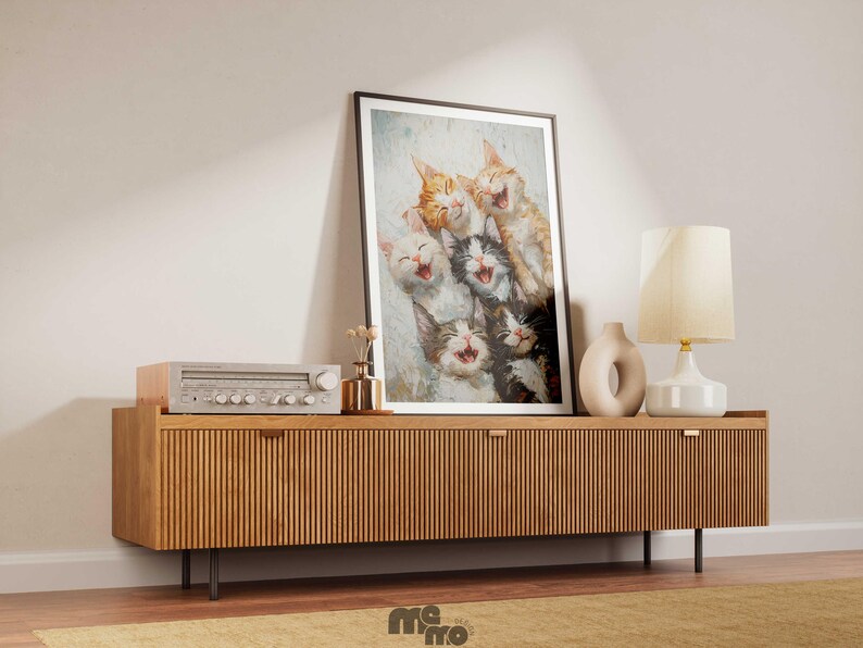 May include: A framed art print featuring a group of laughing cats in various colors, displayed on a wooden cabinet. The cabinet has a ribbed design and is accompanied by a lamp, a vase, and a vintage stereo. The scene is set against a neutral wall.