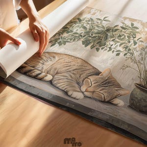 May include: A rolled-up print of a sleeping tabby cat, with a plant in a pot to the right. The cat is lying on a ledge, with a light background. The print is being unrolled on a wooden surface.