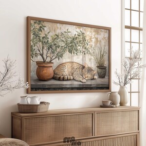 May include: A framed artwork depicts a sleeping tabby cat, potted plants, and a weathered wall. The artwork is in a brown wooden frame. The scene is set on a stone ledge. The artwork is displayed above a wooden cabinet with a woven front.