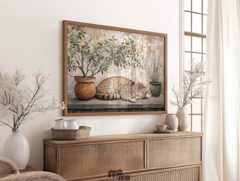 May include: A framed artwork depicts a sleeping tabby cat, potted plants, and a weathered wall. The artwork is in a brown wooden frame. The scene is set on a stone ledge. The artwork is displayed above a wooden cabinet with a woven front.