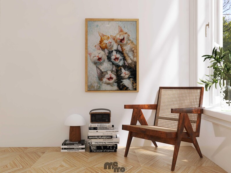 May include: A framed print featuring a group of laughing kittens in various colors, including orange, white, and black. The artwork is displayed on a white wall above a wooden chair and a stack of books.