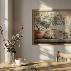 May include: A framed print depicts a sleeping tabby cat, potted plants, and a weathered wall. The artwork is in a wooden frame. A vase of flowers and a cup of coffee on a saucer sit on a wooden table near a window.