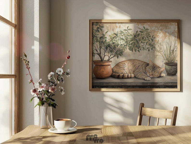 May include: A framed print depicts a sleeping tabby cat, potted plants, and a weathered wall. The artwork is in a wooden frame. A vase of flowers and a cup of coffee on a saucer sit on a wooden table near a window.