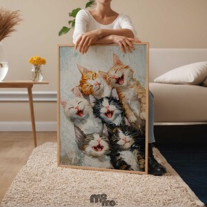 May include: A framed painting featuring six laughing cats in various colors, including orange, white, and black. The artwork has a light blue and white background and is held by a person. The painting is displayed in a room with neutral tones.