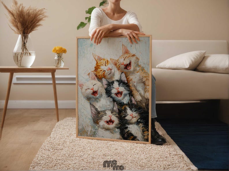 May include: A framed painting featuring six laughing cats in various colors, including orange, white, and black. The artwork has a light blue and white background and is held by a person. The painting is displayed in a room with neutral tones.