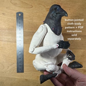 May include: A white cloth body with button joints, posed in a seated position. The body is made of fabric and has a black crow head. The text "button-jointed cloth body pattern + PDF instructions sold separately" is visible in the image.