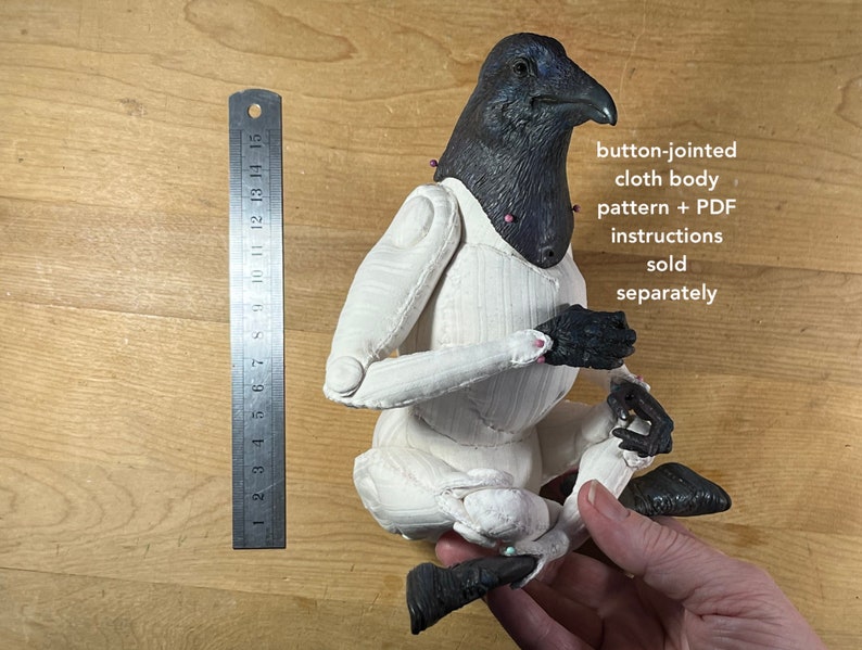 May include: A white cloth body with button joints, posed in a seated position. The body is made of fabric and has a black crow head. The text "button-jointed cloth body pattern + PDF instructions sold separately" is visible in the image.