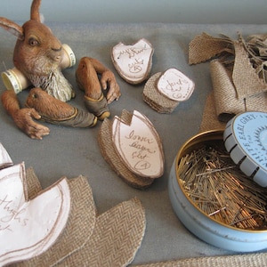 Puede incluir: Una figurita de conejo marrón con un patrón de tela blanca y marrón en su cuerpo. El conejo está rodeado de varios trozos de tela, incluyendo arpillera y un trozo de tela con las palabras "Lower back cut 4" escritas en él. También hay una lata de alfileres.