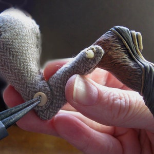 May include: A close-up of a hand holding a brown and gray fabric bird with a button on the body. The bird is being attached to a brown and black sculpted wing with a pair of pliers.