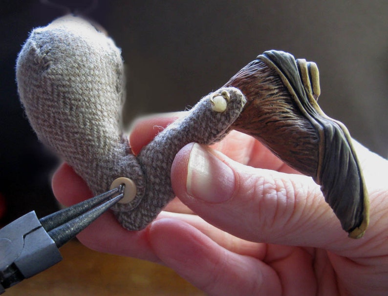 May include: A close-up of a hand holding a brown and gray fabric bird with a button on the body. The bird is being attached to a brown and black sculpted wing with a pair of pliers.
