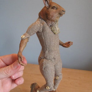 May include: A brown rabbit puppet wearing a tweed suit with button joints. The puppet is posed in a walking position with its right leg forward.