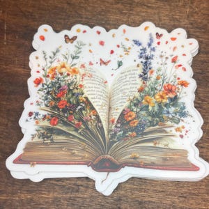 May include: An open book with a red spine and pages filled with text.  Flowers and butterflies are bursting out of the book, creating a whimsical and magical scene.