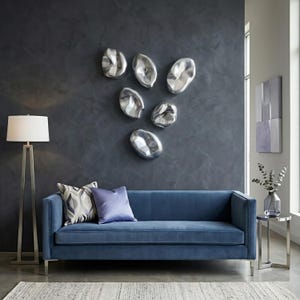 Puede incluir: Un salón moderno con un sofá de terciopelo azul, cojines decorativos, una lámpara de pie plateada y una mesita auxiliar. La pared gris oscuro está adornada con arte mural abstracto plateado. Una alfombra neutra completa el espacio.