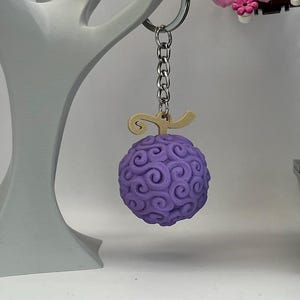 Può includere: Un portachiavi sferico viola con un motivo a spirale, sospeso da una catena d'argento e una chiusura color oro. Il ciondolo è un design dettagliato e tridimensionale. Lo sfondo include una scultura bianca a forma di albero e una composizione floreale decorativa.