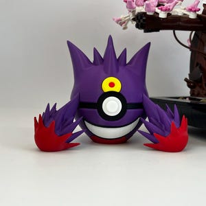 May include: A purple and red Pokemon figure with a white and black sphere design. The figure has a yellow eye and a smiling mouth. The figure is in front of a Lego tree with pink flowers.