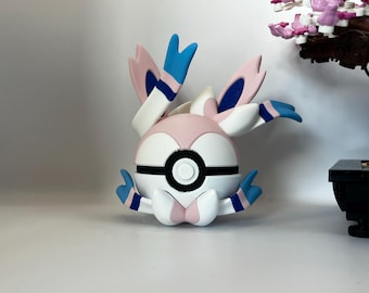 May include: A Sylveon figurine, a Pokémon character, is depicted in a studio shot. The figure is primarily white, pink, and blue, with a round body and large, ear-like appendages. The figure is set against a white background.