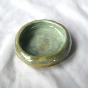May include: A small, round ceramic bowl with a glossy finish. The interior features a blend of soft seafoam green and pale blue tones, while the thick, rounded rim transitions into a warm, earthy golden-yellow hue with subtle colour variations.