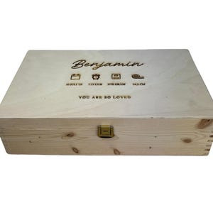 May include: A light-colored wooden box with a hinged lid and a gold-colored clasp. The box is engraved with the name "Benjamin", along with birth details and the phrase "YOU ARE SO LOVED".