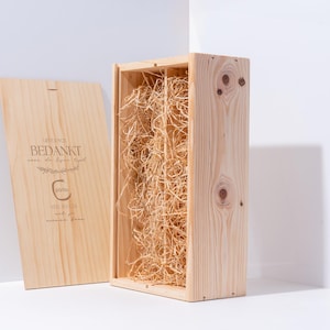 Può includere: Una scatola regalo in legno color chiaro con coperchio, parzialmente aperta per rivelare materiale di imballaggio sminuzzato. Il coperchio ha testo inciso e un logo. La scatola è in legno color chiaro.