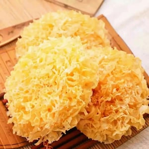 以下が含まれることがあります： 乾燥した薄い黄色の雪キノコ（白キノコまたは銀耳としても知られています）のクローズアップ。 キノコは繊細で縮れた外観をしており、木製の表面に配置されています。