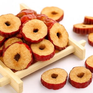 Puede incluir: Dátiles rojos secos, también conocidos como azufaifas, dispuestos en una pequeña bandeja de madera y esparcidos sobre una superficie blanca. Las frutas son de color marrón rojizo con un centro de color beige claro y un agujero en el medio.