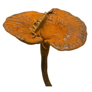 May include: A detailed, close-up view of a mushroom sculpture. The mushroom has a large, textured, orange-brown cap with darker brown accents. A thin, curled, golden-brown element sits atop the cap. The stem is a dark brown color.