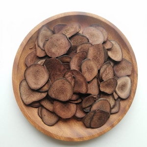 Puede incluir: Un cuenco de madera lleno de numerosas rodajas marrones de forma irregular. Las rodajas tienen una superficie texturizada y diferentes tonos de marrón. El cuenco en sí es de madera clara, creando un contraste con el contenido.