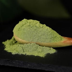 以下が含まれることがあります： 黒い表面にこぼれた緑色の粉末が入った木のスプーンのクローズアップ。粉末は鮮やかな緑色で、スプーンは明るい色の木でできています。背景は暗く、ぼやけています。