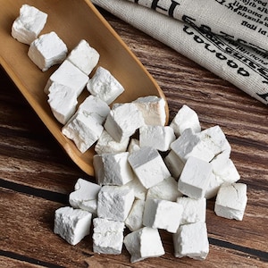 May include: A collection of white, cube-shaped herbal medicine pieces, some spilling from a wooden scoop onto a wooden surface. The pieces have a rough, textured appearance. A folded cloth with text is in the background.