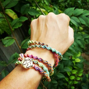 May include: A close-up of a person's wrist wearing a stack of colorful braided bracelets with a silver charm in the shape of a world map.