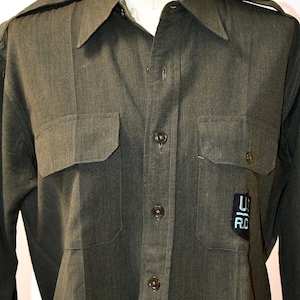 Może przedstawiać: Długorękawna koszula w ciemnozielonym kolorze z zapięciem na guziki z przodu i dwoma kieszeniami z klapkami. Koszula ma naszywkę po prawej stronie z literami "U" i "RO" w kolorze białym.