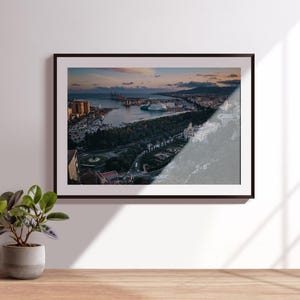 Op de afbeelding: Ingelijste luchtfoto van een kuststad bij zonsondergang. De afbeelding toont een haven met schepen, gebouwen en een park. De foto staat in een zwarte lijst en wordt op een witte muur getoond.