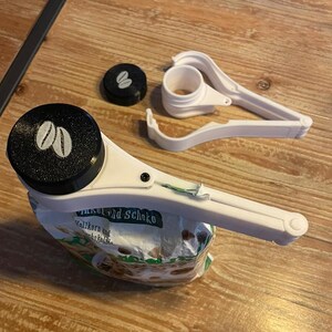 Könnte beinhalten: Weiße Plastik-Beutelclips und ein schwarzer Deckel mit Kaffeebohnen-Design. Der Beutelclip ist an einem Beutel mit Vollkorn und Schoko befestigt. Deckel und Clip liegen auf einer Holzoberfläche.