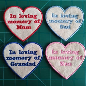 May include: Four heart-shaped patches with embroidered text. Each patch has a white center and a coloured border. The text reads "In loving memory of Mum", "Dad", "Grandad", and "Nan" in red, blue, blue, and pink.