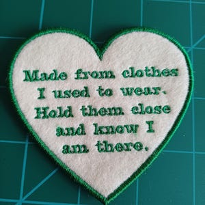May include: A heart-shaped patch with a white centre and green embroidered border. The patch features the text "Made from clothes I used to wear. Hold them close and know I am there" in green lettering.