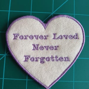 May include: A heart-shaped patch with the embroidered words "Forever Loved Never Forgotten." The patch has a white centre with a purple border and lettering. The background is a green cutting mat.