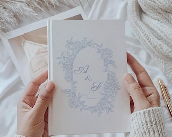 May include: A cream-colored book with a floral design and the initials "A & F" in light blue. The book is held open, revealing a glimpse of a white garment. A gold pen rests nearby on a white surface.