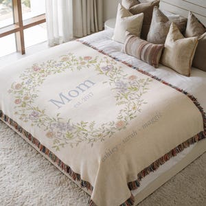 May include: Cream-colored throw blanket with a floral wreath design. The wreath surrounds the word "Mom" and the year "est. 2017". The blanket has a fringed edge and the names "ashley - sarah - maggie" are printed on the bottom.