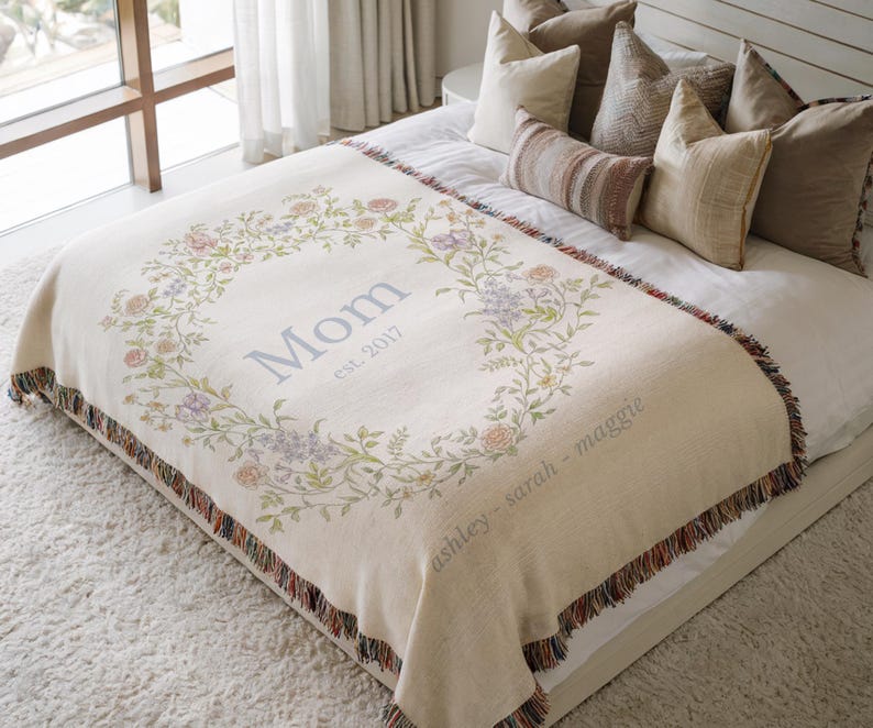 May include: Cream-colored throw blanket with a floral wreath design. The wreath surrounds the word "Mom" and the year "est. 2017". The blanket has a fringed edge and the names "ashley - sarah - maggie" are printed on the bottom.