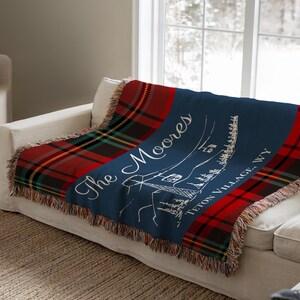 Può includere: Una coperta a quadri blu navy e rossa con le parole "The Moores" e "Teton Village, WY" in corsivo bianco. La coperta ha un bordo con frange ed è drappeggiata su un divano bianco.