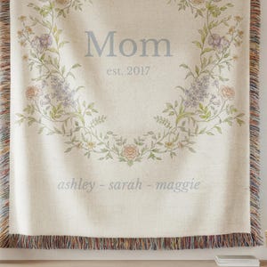 May include: Cream-colored woven throw blanket with a floral wreath design. The word "Mom" and "est. 2017" are centered within the wreath. The names "ashley - sarah - maggie" are below. The blanket has a fringed edge.
