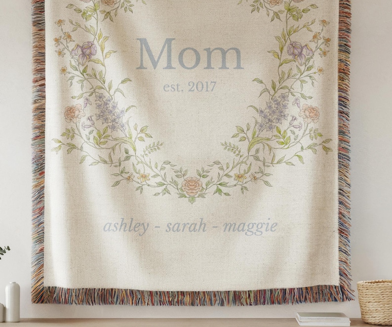 May include: Cream-colored woven throw blanket with a floral wreath design. The word "Mom" and "est. 2017" are centered within the wreath. The names "ashley - sarah - maggie" are below. The blanket has a fringed edge.