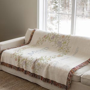 May include: A cream-colored throw blanket draped over a sofa. The blanket features a floral wreath design with the word "Mom" and names. The edges are finished with a fringed trim in shades of brown, red, and orange.