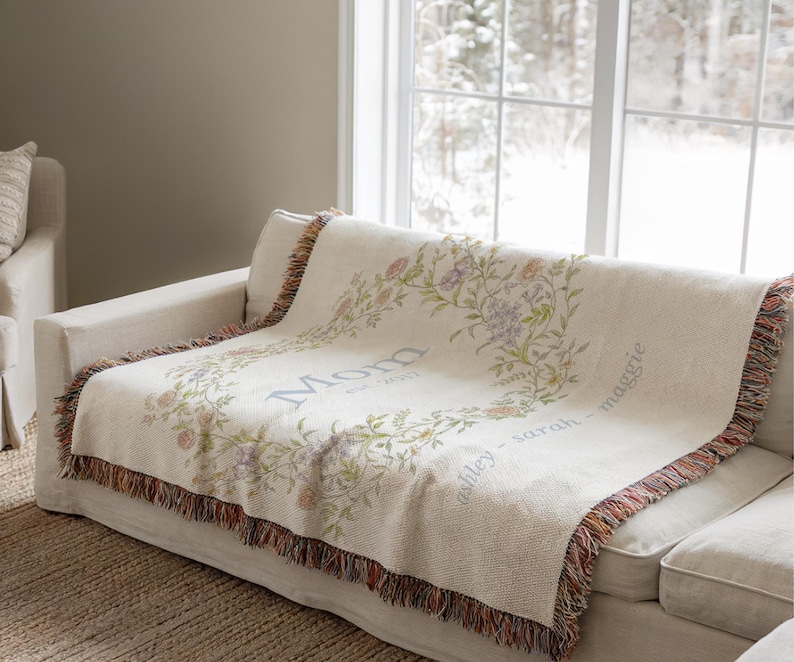 May include: A cream-colored throw blanket draped over a sofa. The blanket features a floral wreath design with the word "Mom" and names. The edges are finished with a fringed trim in shades of brown, red, and orange.