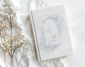 May include: A white wedding guest book with a light blue floral design and the initials "A & F" inside a decorative arch. The date "06.10.2023" is also visible. The book is on a white cloth, with white flowers in the background.