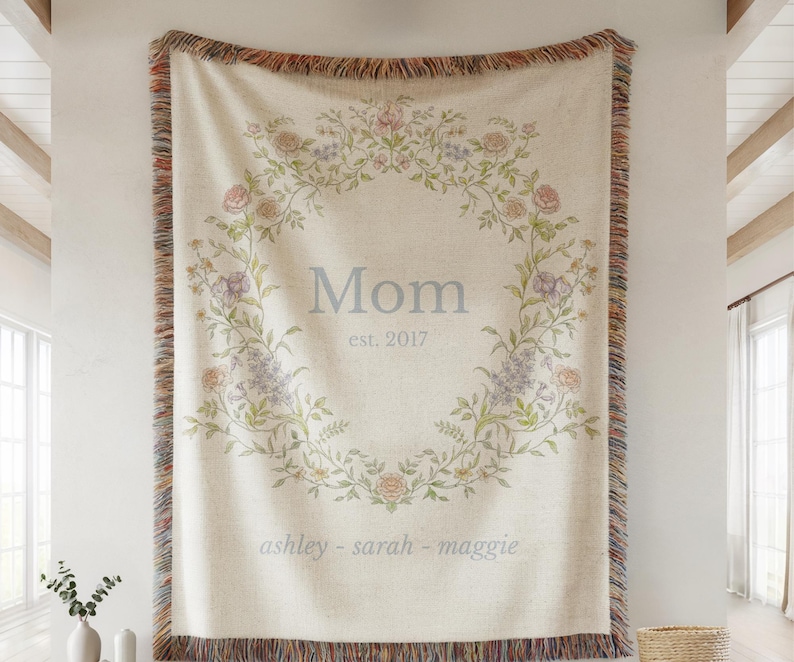 Personalized woven blanket hanging on a wall, featuring the word “Mom” in soft blue text, with “est. 2017” underneath and names “ashley – sarah – maggie” along the bottom. The design is surrounded by a delicate floral wreath in pastel shades of pink, purple, and green, with multicolored fringe along the edges. Displayed in a bright, airy room with white walls and wood ceiling beams.