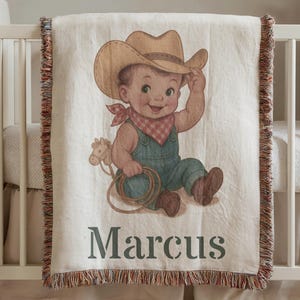 Könnte beinhalten: Eine cremefarbene Babydecke mit einer Illustration zum Thema Cowboy. Das Bild zeigt ein lächelndes Baby mit Cowboyhut und Latzhose, das einen Lasso und ein Spielzeugpferd hält. Der Name "Marcus" ist unten aufgedruckt.