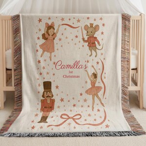 May include: A cream-colored woven blanket featuring illustrations of a ballerina, a nutcracker, a mouse soldier, and a girl. The blanket has a fringe border and the text "Camilla's 1st Christmas" in a script font.