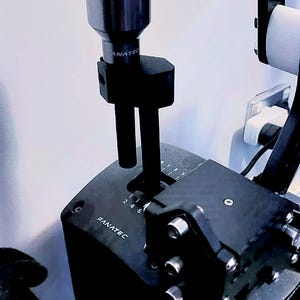 May include: A black Fanatec gear shifter featuring a metallic silver cylindrical knob, a vertical shaft, and a base with gear position markings 1, 2, 3, 4, 5, and 6. The unit includes a black mounting bracket and several small toggle switches labeled TGL.
