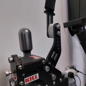 Puede incluir: Un freno de mano de carreras negro con un rodillo blanco y una empuñadura de espuma negra. El freno de mano tiene una placa de fibra de carbono con la palabra "KILL" impresa en ella. El freno de mano está unido a una base de metal con varios tornillos.